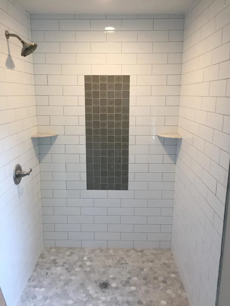 Completed custom shower tile installation featuring white subway tile walls, a vertical grey mosaic accent strip, and a hexagonal marble mosaic floor with corner shelves and fixtures.