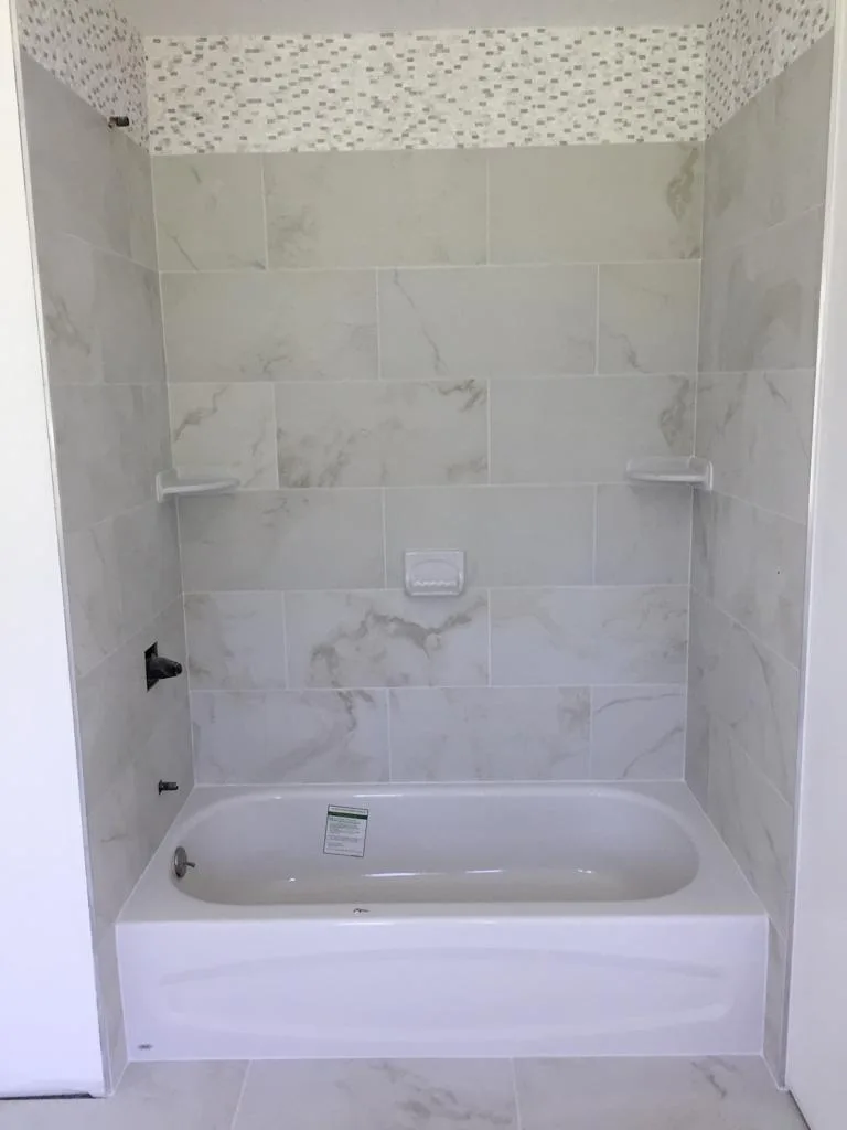 Newly completed bathtub surround tile installation featuring large marble-look wall tiles, integrated corner shelves, and a top mosaic border over a white tub.